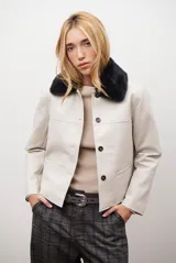 Chaqueta corta color beige con cuello de piel sintética negra, cierre frontal de botones y bolsillos laterales.