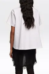 Remera blanca de algodón, de corte línea A con vuelo, escote redondo y mangas cortas. Presenta un estampado frontal con diseño de zapatillas de ballet en tonos grises y lazos rojos.
