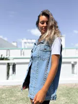Chaleco de jean azul claro con diseño sin mangas, cuello clásico y cierre frontal con botones metálicos. Presenta una profusión de tachas plateadas distribuidas a lo largo de las costuras frontales y los bolsillos del pecho.