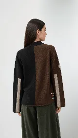 Polera de cuello alto confeccionada en lana lambswool, con diseño bicolor en negro y marrón. Presenta mangas largas con franjas verticales en gris y un parche circular bordado en la manga derecha.
