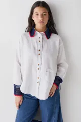 Camisa de lino blanco con cuello y puños tejidos a mano en crochet de color azul con borde rojo. Presenta cierre frontal con botones, bolsillo lateral y costuras a la vista en contraste.