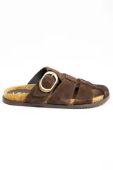 Sandalias tipo zueco de cuero gamuzado color marrón, con diseño de tiras cruzadas, puntera cerrada y una gran hebilla metálica dorada en el lateral. Cuenta con base de corcho y suela plana.