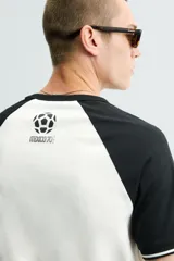 Camiseta de corte regular con diseño color block, cuerpo en color blanco y mangas ranglán en color negro. Presenta cuello redondo, detalles de rib a contraste en los puños y estampado frontal con la leyenda Mexico 70.