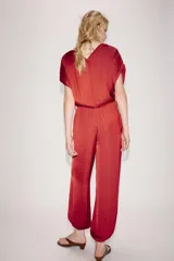 Mono largo satinado de color rojo, con escote en V cruzado y manga corta. Presenta cintura elástica con cinturón negro incorporado, bolsillos laterales ocultos y terminación de las piernas con puño elástico.
