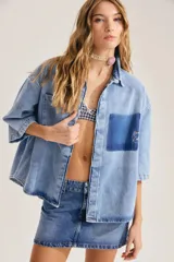 Camisa oversize de denim azul claro, con cuello clásico, mangas cortas a la altura del codo y bolsillo parche en tono más oscuro con detalle bordado en el pecho.