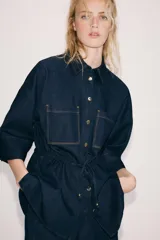 Camisa de denim azul oscuro con cuello solapa, mangas amplias por debajo del codo y bolsillos de parche en el frente. Cuenta con cintura ajustable mediante cordón y cierre frontal con botones metálicos.