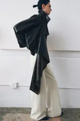 Chaqueta de efecto piel en color negro con cuello subido y bufanda desmontable integrada mediante botones. Presenta manga larga con trabillas ajustables en los puños, cierre frontal con cremallera y bolsillos laterales ocultos en las costuras. El diseño incluye un bajo con elástico en la espalda y forro interior.