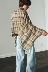 Capa corta con estampado a cuadros en tonos mostaza, marrón y blanco. Presenta un cuello redondo y mangas amplias tipo murciélago. Incluye un lazo o foulard integrado en el cuello para anudar.