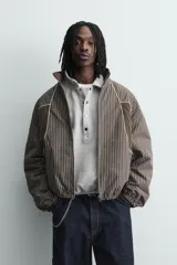 Cazadora de corte boxy fit confeccionada en tejido técnico. Presenta un diseño a rayas verticales, cuello subido, cierre frontal de cremallera y mangas largas con puños elásticos. Incluye bolsillos laterales con cremallera oculta y detalles de paneles en contraste.