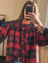 Camisa de franela a cuadros rojos y negros, con cuello redondo, volados decorativos en la parte frontal y mangas largas abullonadas con puños elásticos.