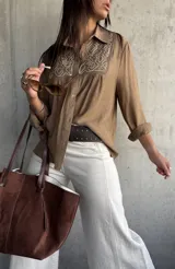 Camisa de color camel con cuello clásico, manga larga y detalle de bordado geométrico en color blanco en la parte frontal superior. Presenta cierre frontal con botones y un corte holgado.