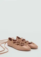 Zapato plano tipo chatita color nude, confeccionado en cuero, con diseño de tiras cruzadas y cordones para atar alrededor del tobillo, ojalillos metálicos dorados y punta redonda.