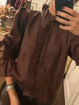 Camisa marrón de cuello alto con volados, mangas abullonadas y puños elásticos fruncidos. Cuenta con una tira de volados en la parte frontal.
