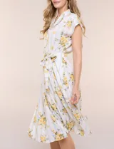 Vestido camisero midi de silueta evasé, confeccionado en tejido liviano blanco con estampado floral amarillo y rayas celestes. Cuenta con cuello solapa en V, cartera con botones frontales, mangas cortas japonesas y cintura definida con lazo ajustable a juego.