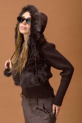 Campera corta de diseño combinado en color marrón. Presenta cuerpo de piel sintética con capucha, mangas tejidas en punto acanalado y cierre frontal con vista de cuero sintético. Incluye detalles de pompones laterales en el ruedo.