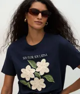 Remera de algodón color blanco, con cuello redondo y manga corta. Presenta un estampado con la frase 'Soy flor. Soy latina' y flores bordadas en relieve con textura tejida.