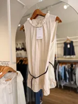 Blusa sin mangas de color beige, confeccionada en mezcla de fibra regenerada y algodón. Presenta un delicado cuello con detalle trenzado, volados sutiles en el ruedo y viene acompañada por un cinturón fino de cordón trenzado oscuro.