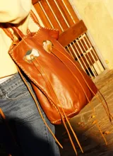Cartera de cuero color camel, de silueta amplia y blanda, con manijas largas y terminación artesanal en los bordes. Cuenta con apliques decorativos frontales de alpaca y nácar, tientos largos colgantes y bolsillo interno.