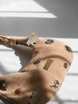 Cartera tipo baguette de gamuza color tostado, con diseño decorativo que incluye diversos apliques metálicos, botones y dijes con formas variadas. Cuenta con una placa metálica con el logo de la marca y cierre superior.