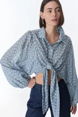 Top de manga larga con diseño de camisa corta, confeccionado en tela celeste con bordado floral blanco. Presenta cuello clásico, cierre frontal con botones y detalle de nudo en la cintura.