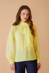 Blusa de gasa color amarillo pastel, con cuello alto y lazo frontal tipo corbatín. Presenta mangas largas con puños abotonados y un diseño elegante y fluido.