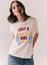 Remera blanca de manga corta y cuello redondo, con estampado frontal de estilo retro con la inscripción "Just a Small Town Girl Journey" acompañada de gráficos florales en tonos rojo, amarillo y azul.