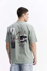 Remera de manga corta color verde salvia, con cuello redondo y un estampado gráfico en la espalda que incluye tipografía en rojo y blanco, y fotografías en blanco y negro.