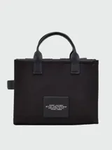 Cartera tote de lona color negro, modelo The Cargo Canvas Medium Tote Bag de Marc Jacobs. Presenta múltiples bolsillos frontales con solapa y cierre de cremallera, asas de mano superiores y una correa de hombro ajustable y extraíble. Cuenta con el nombre de la marca y la inscripción "THE TOTE BAG" estampados en el frente.