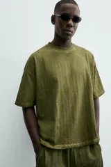 Camiseta de algodón de corte relajado (relaxed fit) con cuello redondo y manga corta. Presenta un tejido acanalado vertical (canalé) y un acabado ligeramente desgastado o lavado en tono verde oliva.