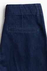 Pantalón de jean tipo wide leg de tiro alto, confeccionado en denim rígido de color azul oscuro. Presenta un corte holgado desde el muslo hasta el ruedo, con raya marcada en el centro de las piernas, cierre con botón oculto y bolsillos al bies.