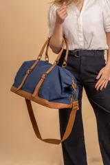 Bolso de viaje amplio confeccionado en tela canvas azul con detalles, base, asas y correas en símil cuero tostado. Cuenta con correas ajustables y herrajes metálicos.