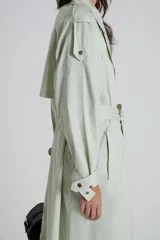 Trench color verde claro con cuello solapa, doble botonadura, cinturón y bolsillos laterales.
