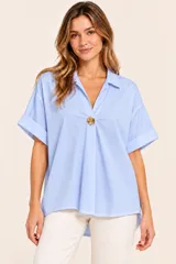 Camisa de algodón celeste, de corte recto, con cuello camisero, escote en V y un botón dorado decorativo en el centro. Cuenta con mangas cortas con vuelta y diseño holgado.