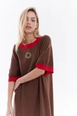 Vestido midi de punto color marrón con calce relajado, cuello redondo, mangas cortas y terminaciones en color rojo contrastante. Cuenta con un diseño bordado en el pecho en forma de sol y detalles en las mangas.