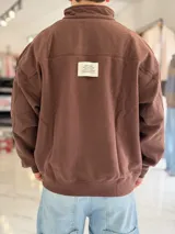 Buzo gris oscuro de corte oversize con cuello alto y cierre frontal parcial. Cuenta con mangas largas y puños elásticos, además de un pequeño bordado con texto en el pecho.