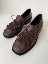 Mocasín de cuero marrón con textura granulada, diseño de horma cuadrada, detalle de flecos y lazo decorativo en el empeine, montado sobre una suela y taco bajo de color negro.