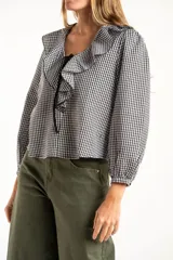 Blusa de manga larga con estampado de cuadros vichy en blanco y negro. Presenta un cuello con volados amplios y un detalle de lazo negro en el centro del escote.