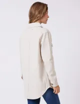 Sobrecamisa de corte amplio confeccionada en tejido suave al tacto, color beige. Presenta cuello de solapa, cierre frontal con botones efecto carey, dos bolsillos frontales con solapa y mangas largas con puños abotonados. El ruedo es redondeado en los laterales y cuenta con canesú en la espalda.