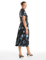 Vestido midi de seda fría con estampado floral en tonos azules sobre fondo negro. Presenta escote en V con cierre frontal de botones, mangas cortas con terminación fruncida, corte en la cintura con lazo de ajuste y falda plisada.