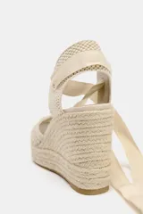 Alpargata de mujer con plataforma y taco cuña alto, confeccionada en rafia trenzada. La capellada es de tejido de punto calado color beige, con talón cerrado y tiras largas de tela para atar al tobillo.