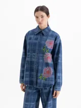 Camisa de denim con diseño a cuadros en tonos azules, cuello clásico, mangas largas y cierre frontal con broches a presión. Presenta bordados florales en el frente y contornos de flores bordados en hilo fino.