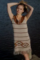 Vestido corto tejido en hilado de algodón color beige con rayas horizontales marrones, cuello de cuero marrón y terminación con flecos. Presenta un diseño geométrico en la parte inferior.