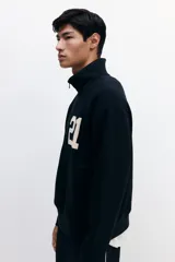 Buzo negro de tejido pesado con interior cepillado suave, cuello elevado con medio cierre y hombros ligeramente caídos. Presenta un estampado frontal con el número "21" en color beige. Puños y bajo en punto acanalado.