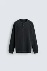 Camiseta de manga larga tipo ranglán con cuello redondo y cierre frontal de botones. Presenta un diseño de rayas horizontales finas en tonos oscuros.