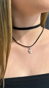 Gargantilla doble de estilo choker, compuesta por una cinta de gamuza negra y un cordón fino negro con un dije metálico plateado en forma de luna con calados de estrella y corazón.