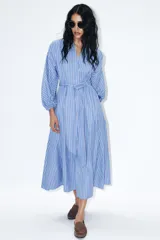 Vestido midi de corte camisero con estampado de rayas verticales en tonos azul, blanco y rojo. Presenta cuello redondo con escote en pico, mangas largas abullonadas con puños elásticos y cinturón ajustable del mismo tejido. Incluye bolsillos laterales ocultos y cierre frontal cruzado con botones.