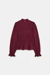 Camisa de manga larga color bordó con cuello subido y detalle de nido de abeja elástico en el pecho. Presenta volantes en el cuello, puños y ruedo, con cierre de botón oculto en la espalda.