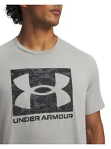 Remera deportiva de manga corta, color gris claro, con cuello redondo y un gran estampado frontal del logo de Under Armour dentro de un recuadro con patrón de camuflaje digital en tonos grises oscuros.