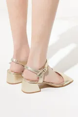 Sandalias doradas con taco cuadrado, tira ancha sobre los dedos y pulsera al tobillo con hebilla.
