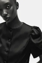 Blusa corta de sarga negra con leve brillo, ajuste entallado, escote redondo, botones forrados al frente y mangas cortas abullonadas con pliegues. Presenta costuras princesa al frente para estilizar la silueta.